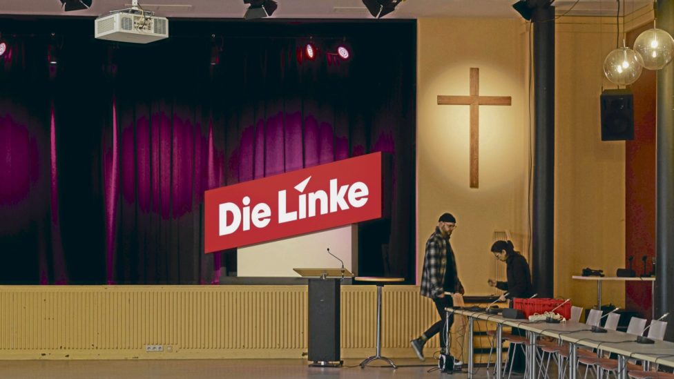 im noch leeren Veranstaltungsraum mit dem Logo "Die Linke" und christlichem Kreuz